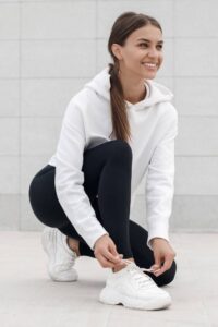 A happy woman in a white hoodie, black leggings, and white trainers ties her shoe lace.