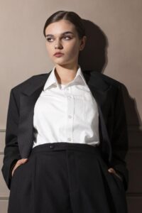 A lady wearing a black suit and tailored white shirt looks into the distance with hands in pockets