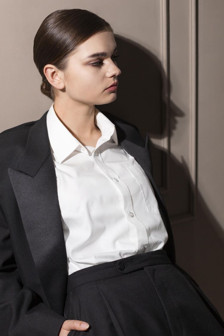 Lady in tailored black suit and white shirt leans against a wall with hands in pockets