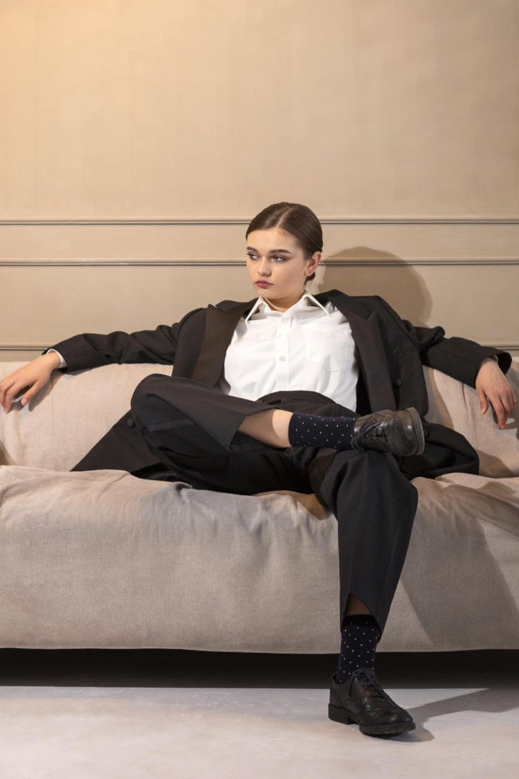 Lady in tailored black suit, white shirt and black shiny shoes sits confidently on a beige sofa