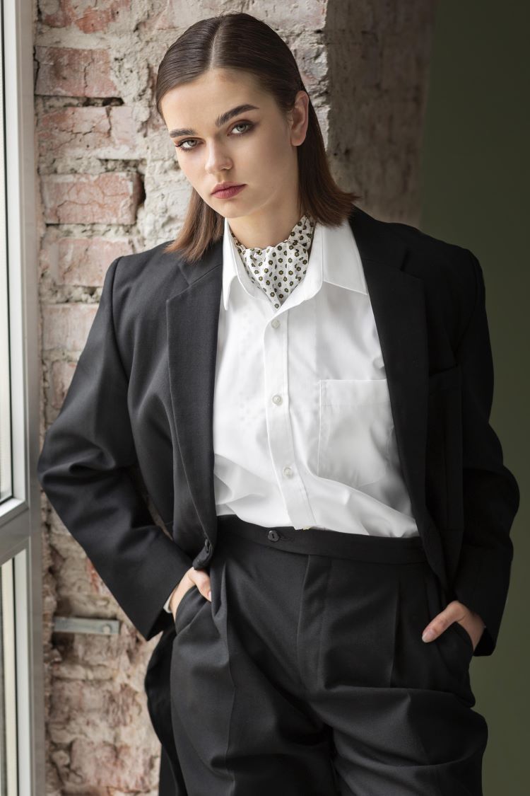 Lady in tailored black suit, white shirt and polka dot silk scarf leans against an indoor brick wall