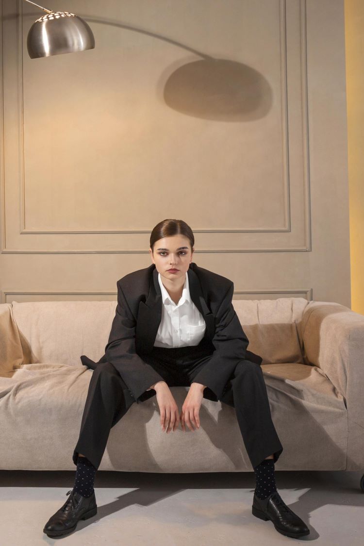 Lady in tailored black suit, white shirt and black shiny shoes sits confidently on a beige sofa