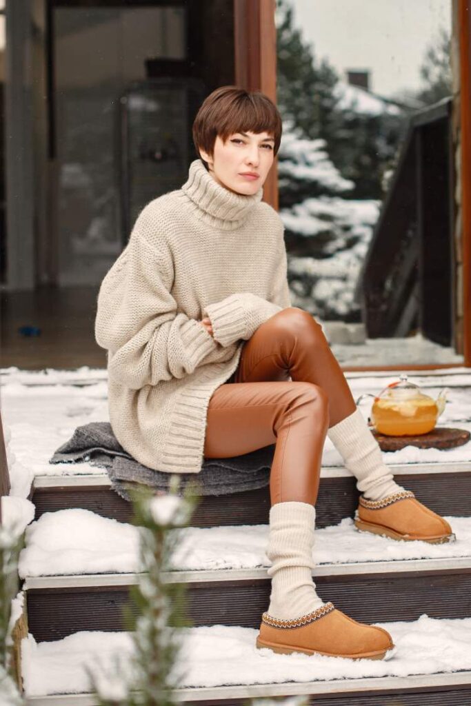 A lady sits on a blanket on the snow-covered steps outside her house. She wears a warm oversized cream jumper with a high neck, long socks to match and warm suede house shoes.