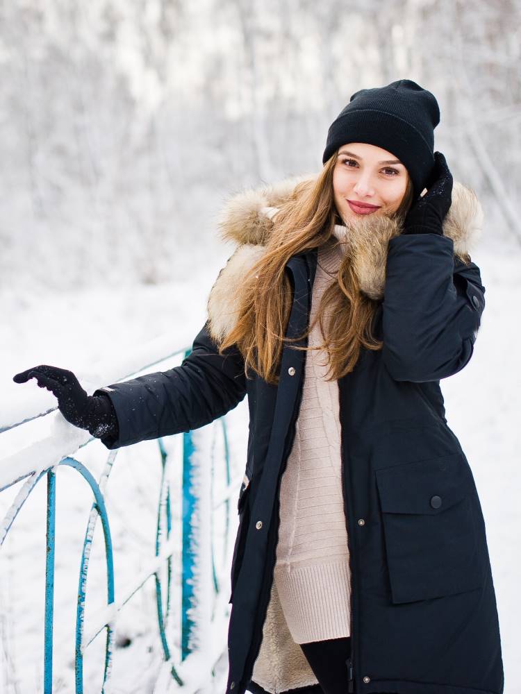 A lady wears a thick black padded jacket over a knit dress to keep warm in a snow covered park.