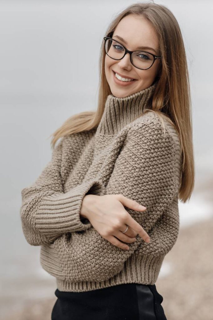 A lady stands on a pebble beach wearing a pale brown waffled woolen jumper.
