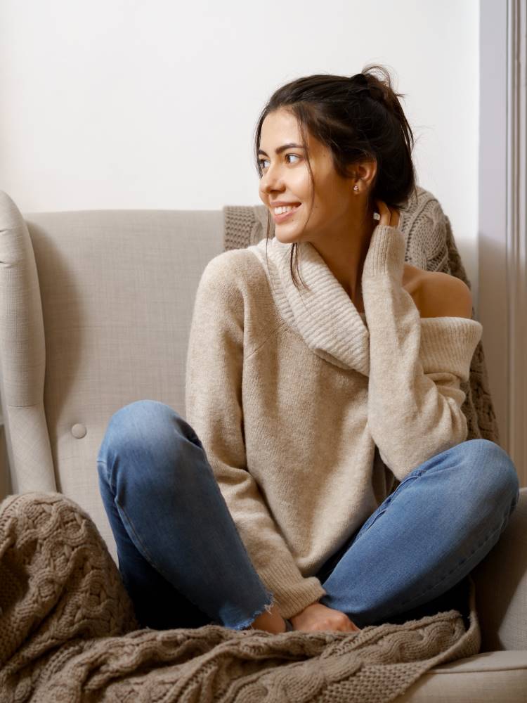 A lady sits cross-legged on an armchair. She wears a cream cashmere jumper with a high loose neck and sits on a warm knitted throw.