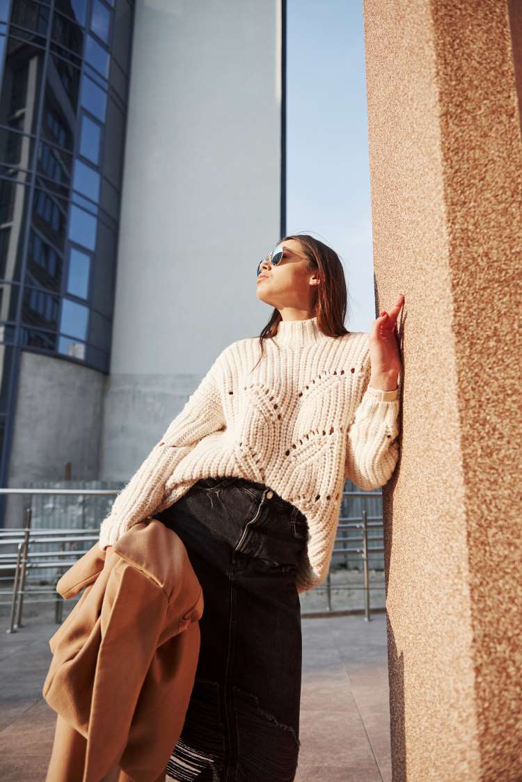 A woman leans against a city office wall in the autumn sun wearing a loose fitting high-necked knitted jumper, black denim skirt and sunglasses.