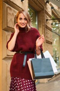 A woman wearing a long red jumper accessorised with a black belt and carrying a few bags from the high-end stores in the street behind her.