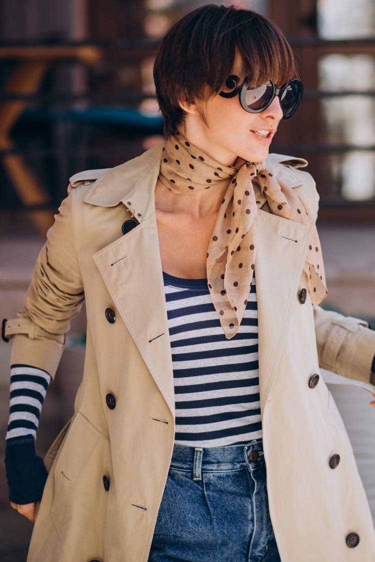 A woman wears a blue-spotted scarf with a cream background, a breton-style blue and white striped top under a trench coat.