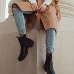 Sitting casually on the plinth of a column, wearing a mid-length brown coat, skinny ripped jeans and big black boots.