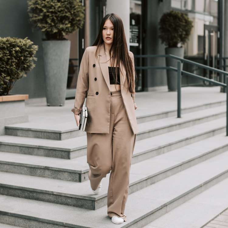 A professional woman wearing a baggy suit and white trainers stands on the steps of an office building.