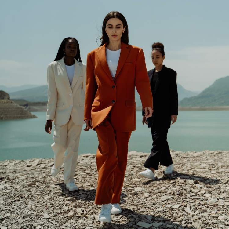 Three women walk purposefully towards the camera across a shingle beach while wearing baggy suits and white trainers. One suit is cream, the next a warm orange and the third suit is black. All give a professional, stylish, and very current vibe.