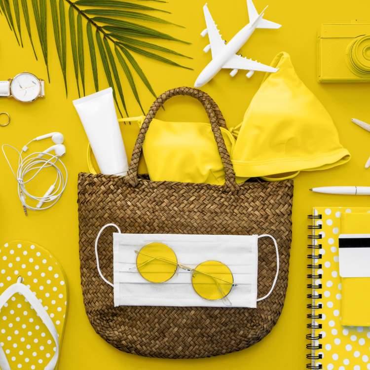 An exploded photo of a beach bag with some essential items to pack: watch, headphones, flip flops, singlasses, mask, bikini, suncream, camera, notebook and pen.