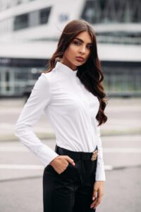 A lady wearing a crisp white shirt and fitted black trousers stands with one hand in her pocket. Her hair is long and neatly brushed, matching the businesslike look of her outfit.