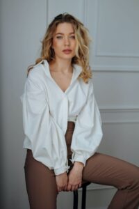 A lady sits on a high stool wearing a balloon-sleeved white shirt and skinny brown trousers.