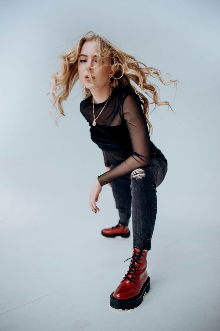 Crouching woman with long blond hair wearing black chiffon top with ripped jeans and red chunky boots