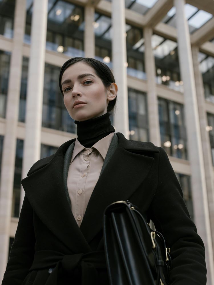 alt="A woman in a fitted black long coat over a grey-beige shirt which is in turn over a black high-necked polo neck, carrying a briefcase."