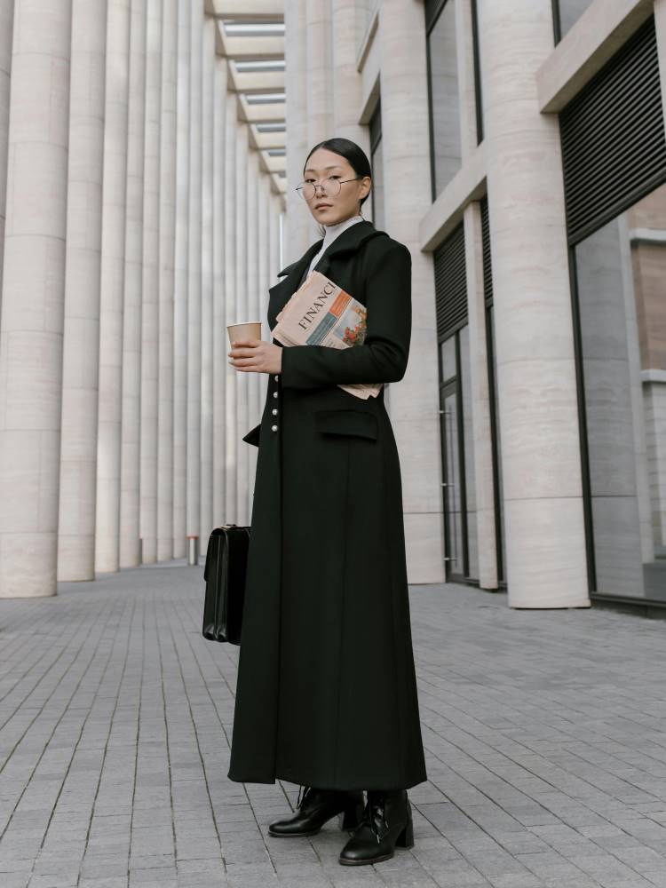 Woman in a long black fitted coat holding a briefcase, newspaper, and coffee to go.