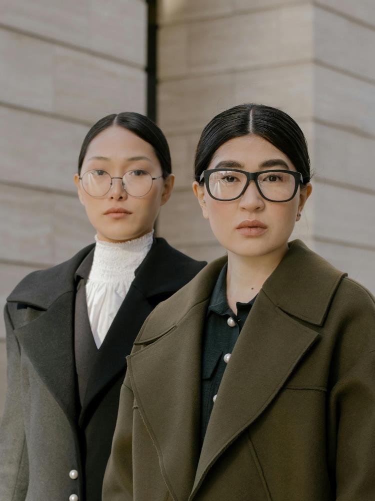 Two women outside their office. One in a long olive coat and black shirt with rounded pearl buttons, the other in a long grey coat with black lapel detail over a high-necked frilly shirt.