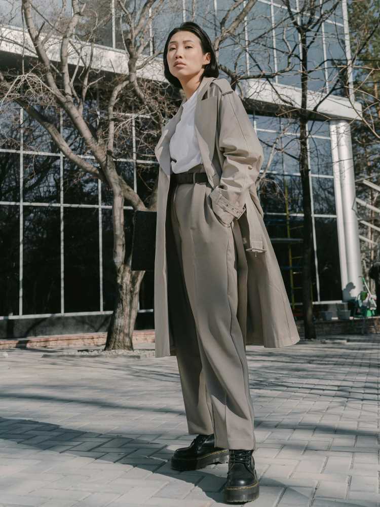 A woman smartly dressed in a light grey trench coat over colour-matched light grey suit trousers and a white shirt.