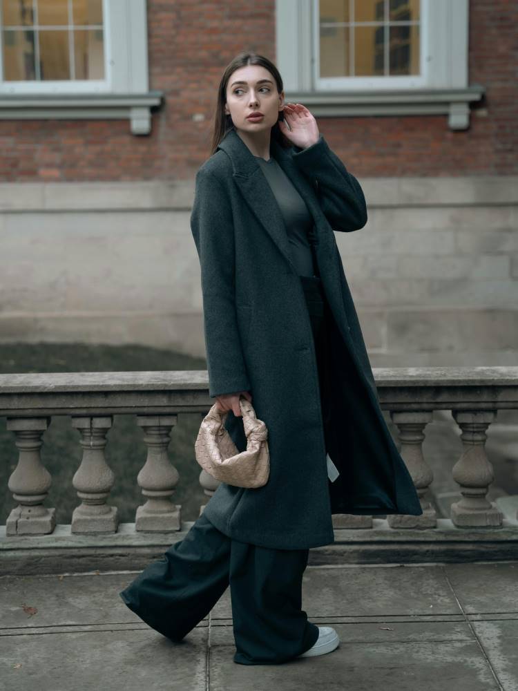 A lady stylishly dressed in baggy comfortable but tailored wear. She wears a long charcoal coat, tailored baggy charcoal trousers, a grey t-shirt, and white trainers.