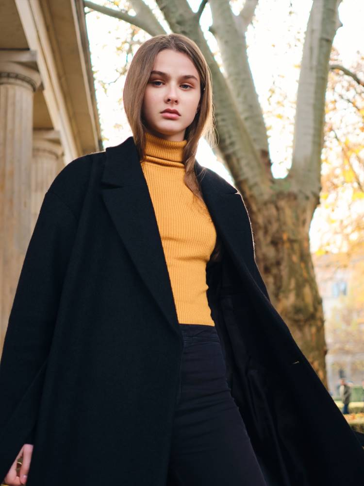 alt="A lady in an oversized but fairly formal long black coat, a pastel orange polo neck and black jeans stands near an old building in a park."